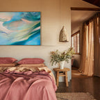 Bedroom with original abstract oil painting, pink bedding, and wooden stool with plants.